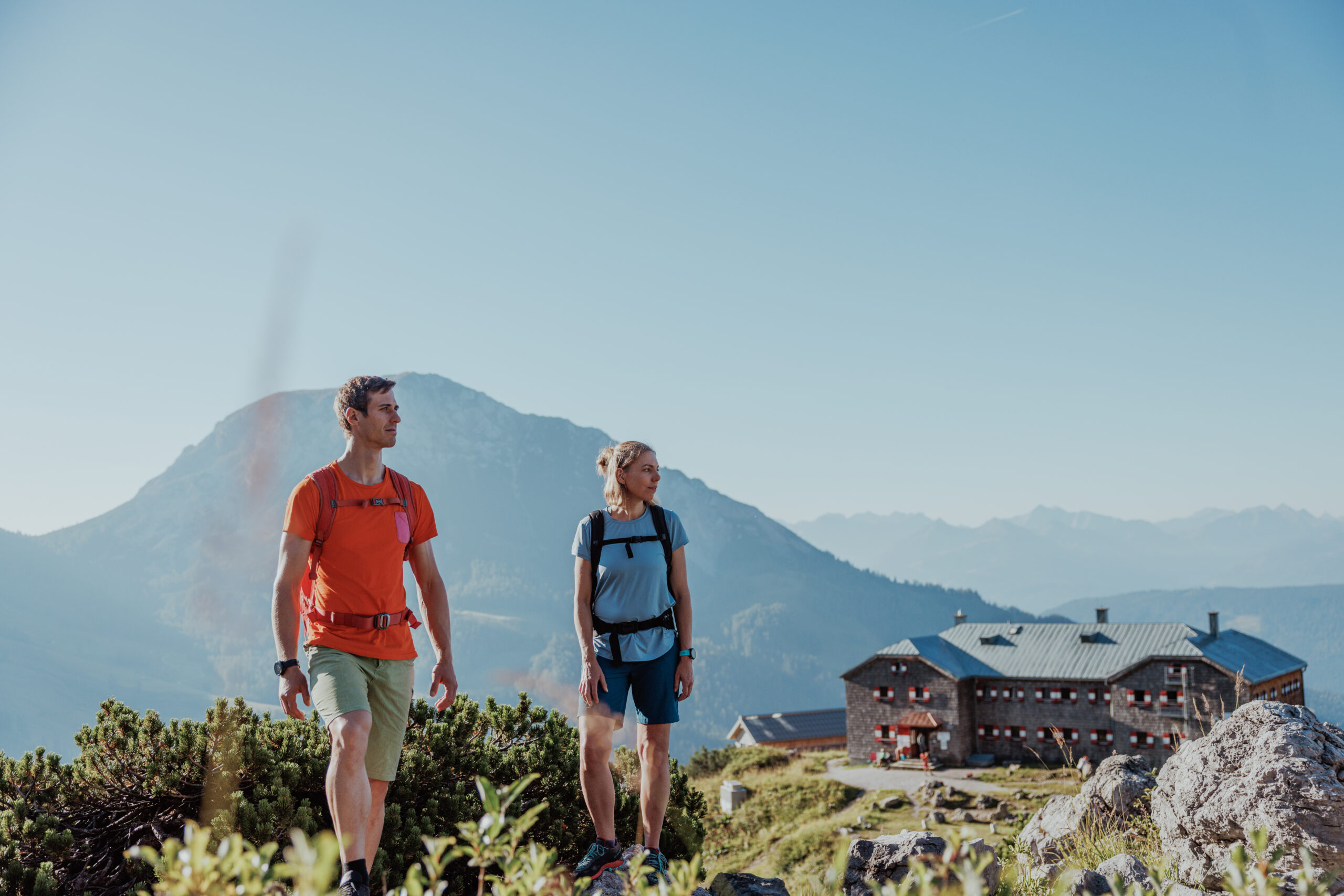 Ein Mann und eine Frau wandern auf einem Wiesenpfad, umgeben von gr&uuml;nen Almfl&auml;chen und gelben Blumen. Hinter ihnen erhebt sich eine markante Alpen-Bergkette unter einem hellen, wolkigen Himmel.