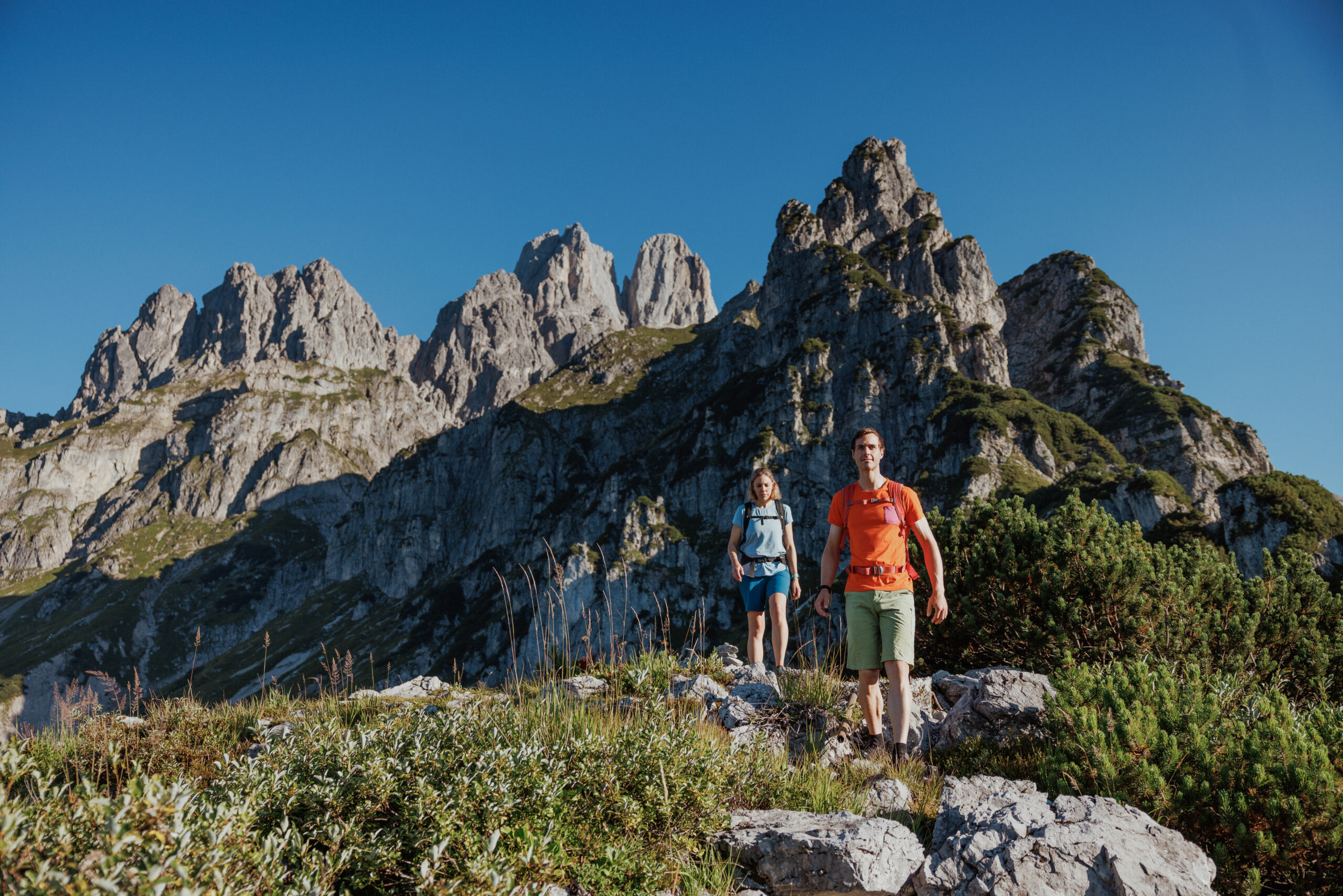 Zwei Wandernde stehen auf einem felsigen Aussichtspunkt vor steilen, gezackten Bergspitzen. Der Himmel ist tiefblau und klar.