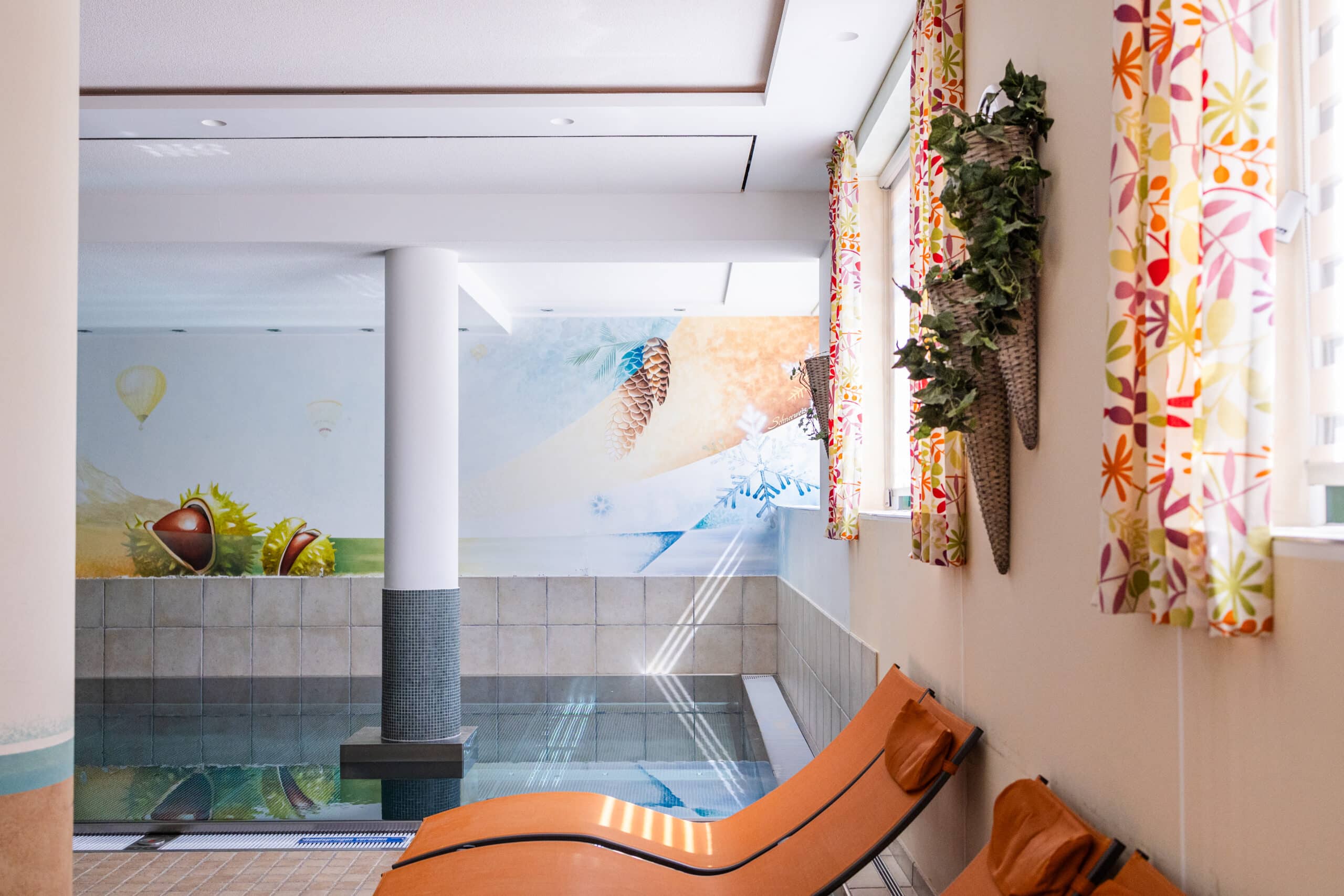 Innenbereich eines Wellnessbades mit Liegest&uuml;hlen und einem kleinen Becken, dekoriert mit bunten Wandmalereien und floralen Vorh&auml;ngen.