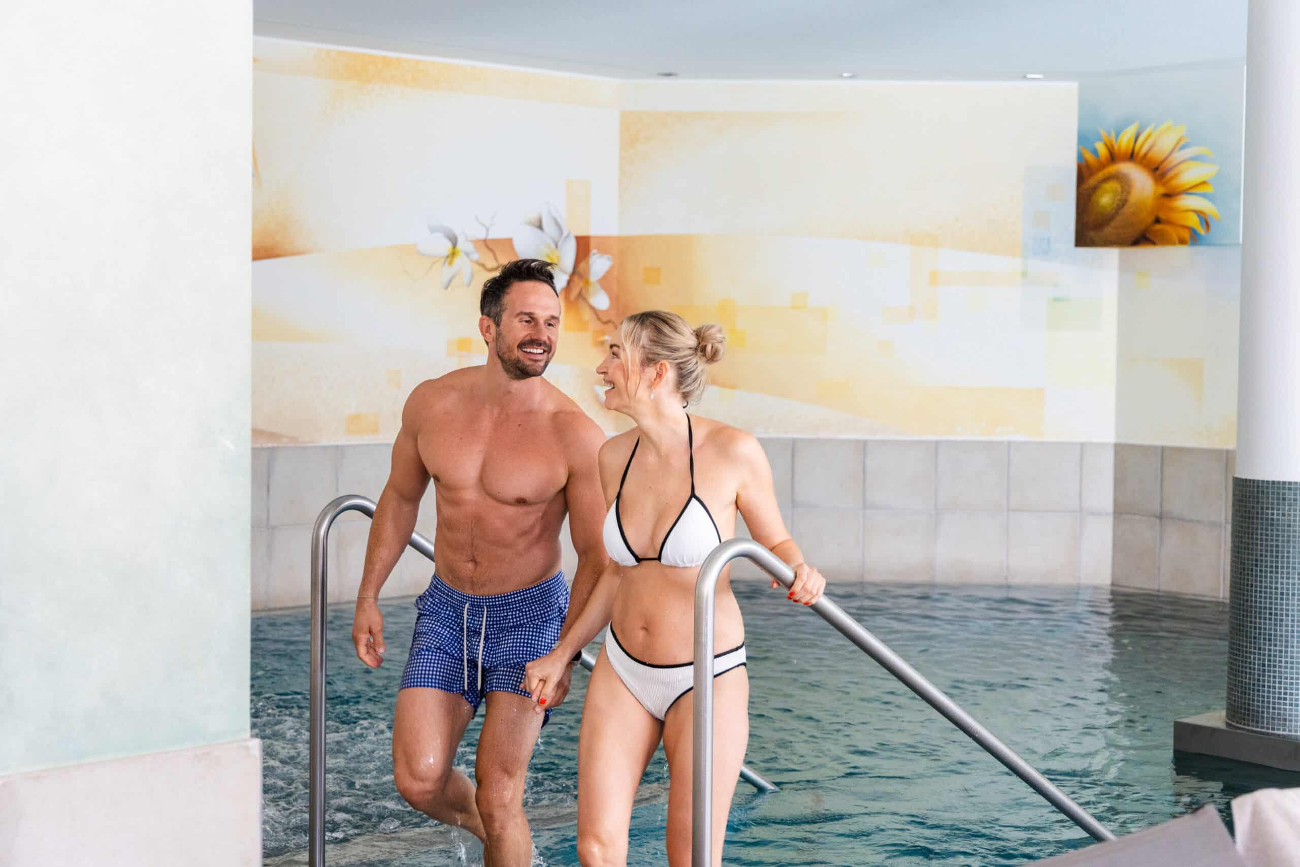Zwei Personen steigen gemeinsam in ein warmes Innenbecken, umgeben von hellen Wandmalereien und Spa-Atmosph&auml;re.